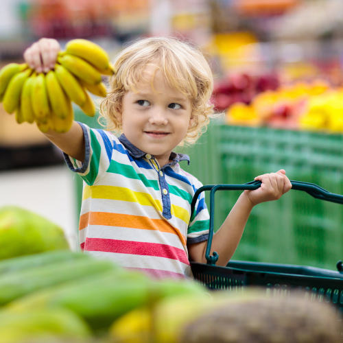 Kid holding up bananas to someone off camera inside a grocery story while holding a grocery basket