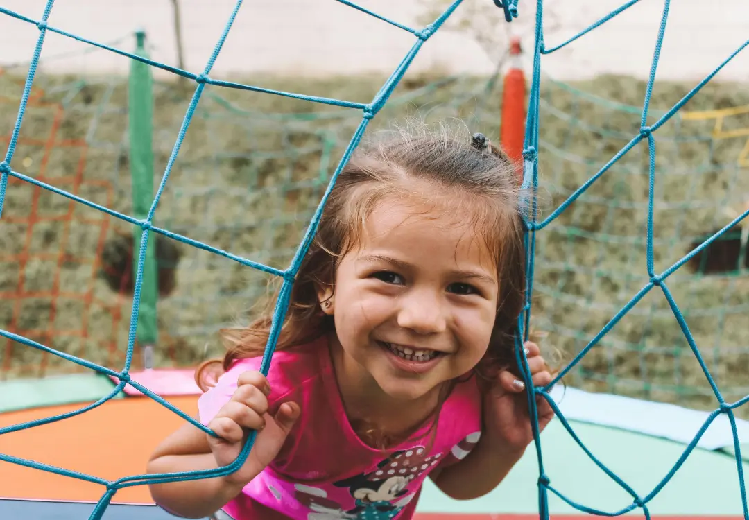 Young girl on a playground smiling at the camera holding onto a climbing net