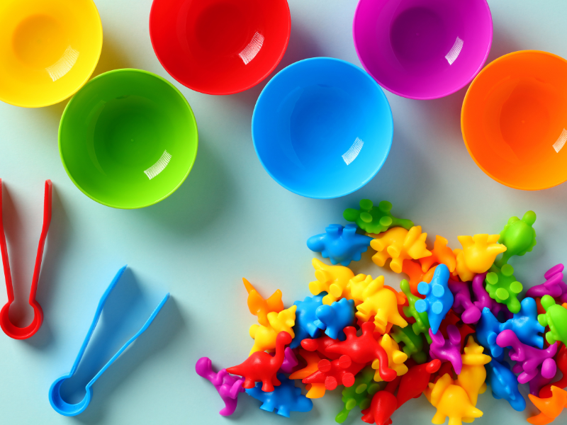 Small colorful plastic dinosaurs in a pile next to small plastic bowls with tongs in different colors, ready to sort